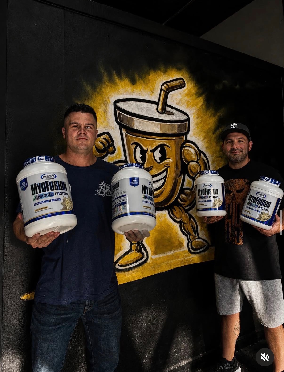 Muscle Lounge founders holding protein tubs in front of the mascot mural